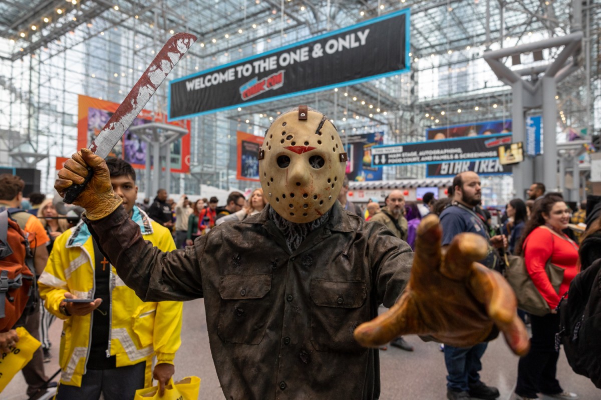 A person dressed as Jason Voorhees from the movie “Friday the 13th” holds up a prop knife in one hand and reaches out to the camera with their other hand.