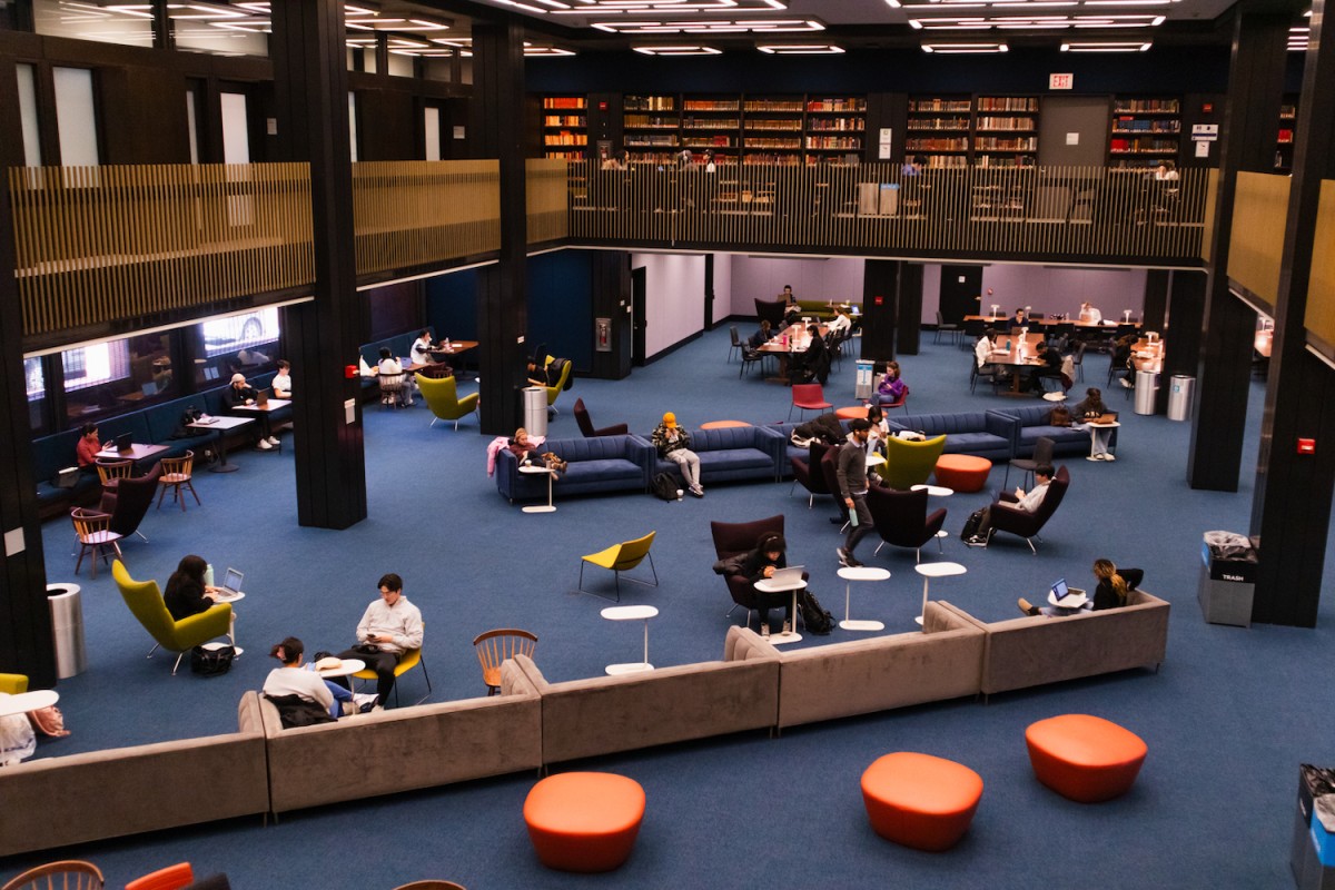 A large room with blue carpeting and a variety of colorful couches and chairs. There are students sitting with their laptops. On the floor above there are bookshelves filled with books.