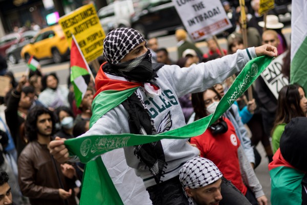 A man in a white hoodie wearing a keffiyeh on his head is standing on another man’s shoulders with an unfurled green banner.