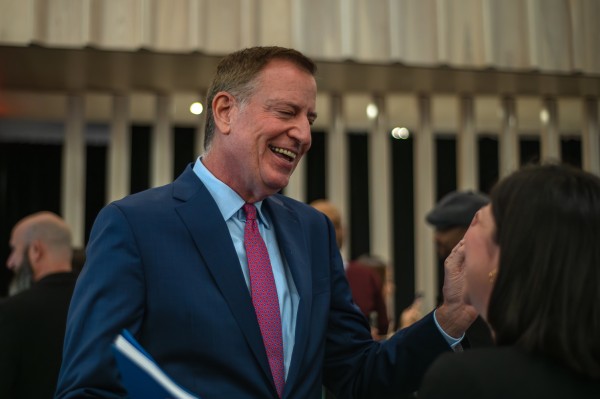 Bill de Blasio laughing and wearing a navy suit, blue shirt and red tie.