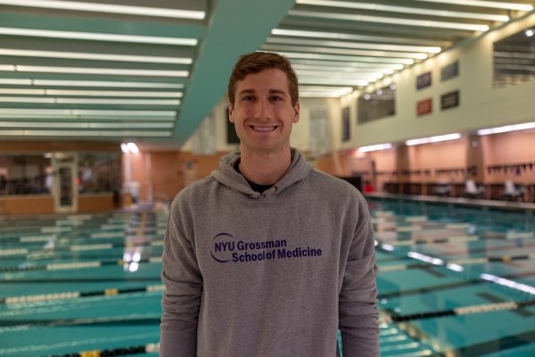 A man is standing in front of a swimming pool and smiling. He is wearing a gray hoodie that reads, “NYU Grossman School of Medicine.”