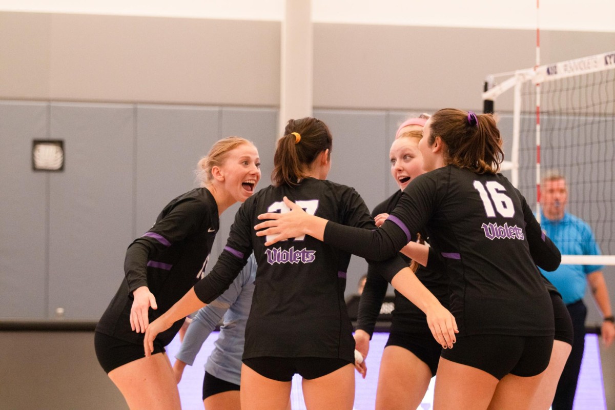 Four athletes are standing and looking at each other with their arms around one another. They are wearing black shirts with ‘Violets’ written in purple on the back. There is a volleyball net to their right.