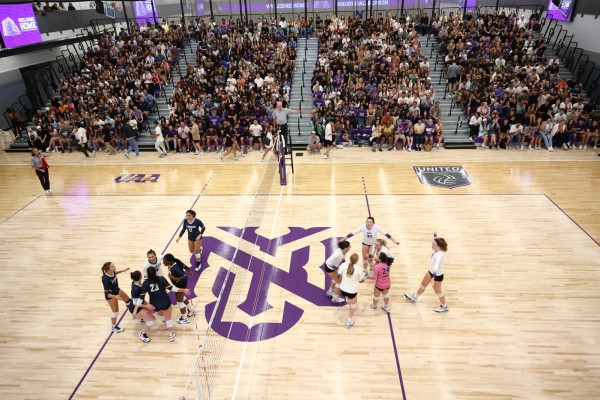 A wide shot of a volleyball court. The team on the left side of the net are wearing shirts with ‘ITHACA’ written on them and the team on the right are wearing shirts that have ‘VIOLETS’ on them. There is a purple N.Y.U. logo on the court and a full crowd.