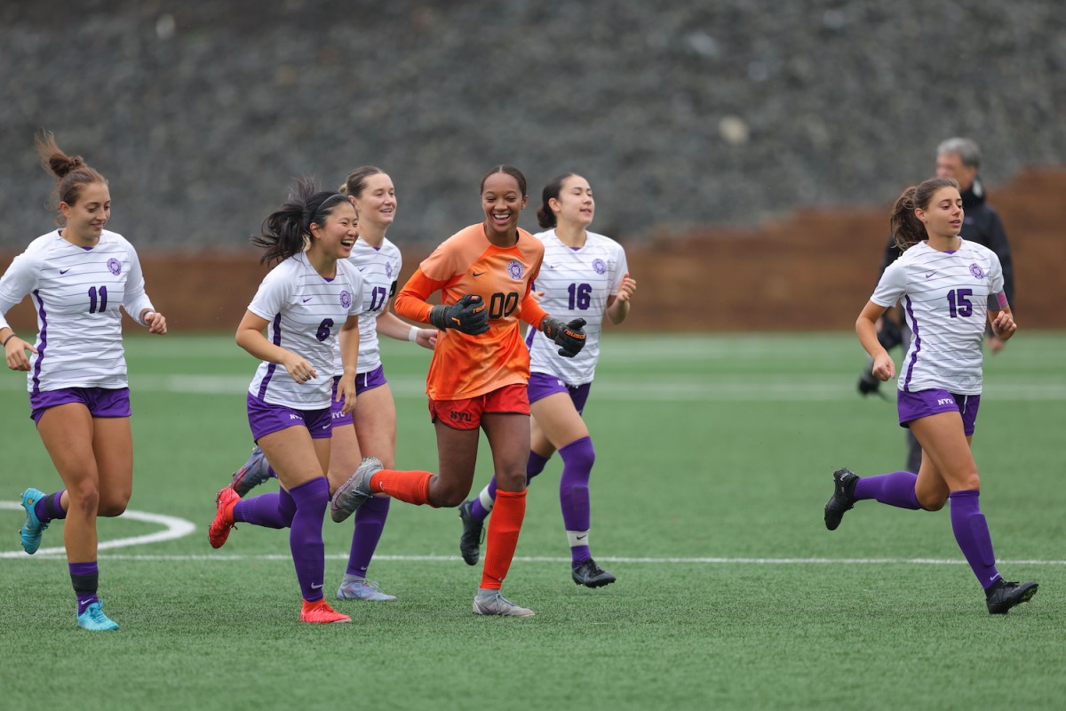Six+soccer+players+are+running+on+a+soccer+pitch+and+smiling.+Five+of+them+are+wearing+white+and+purple+soccer+uniforms+while+one+is+wearing+an+orange+uniform+and+goalkeeper+gloves.