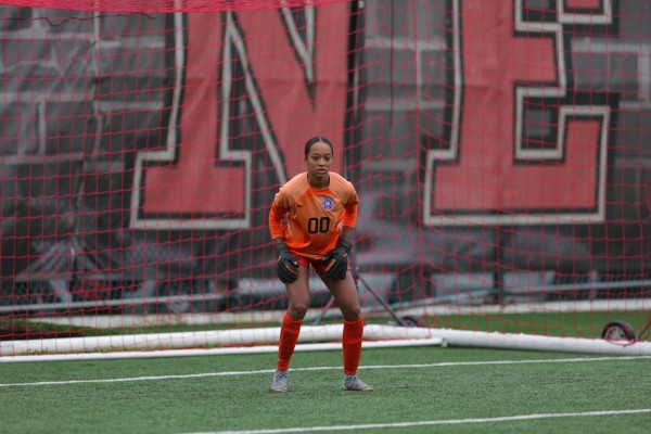 A woman is standing in front of a soccer goal net. She is wearing an red soccer uniform, orange socks, and goalkeeper gloves. Her uniform has the number 00.
