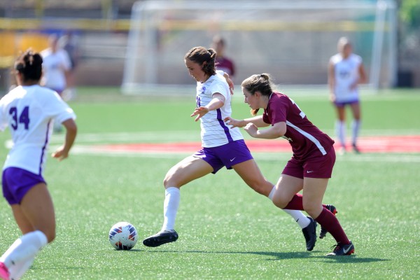 Two soccer players are running after a ball on a soccer pitch. One of them is wearing a white and purple outfit while the other is wearing a red one. There is another player to the left.