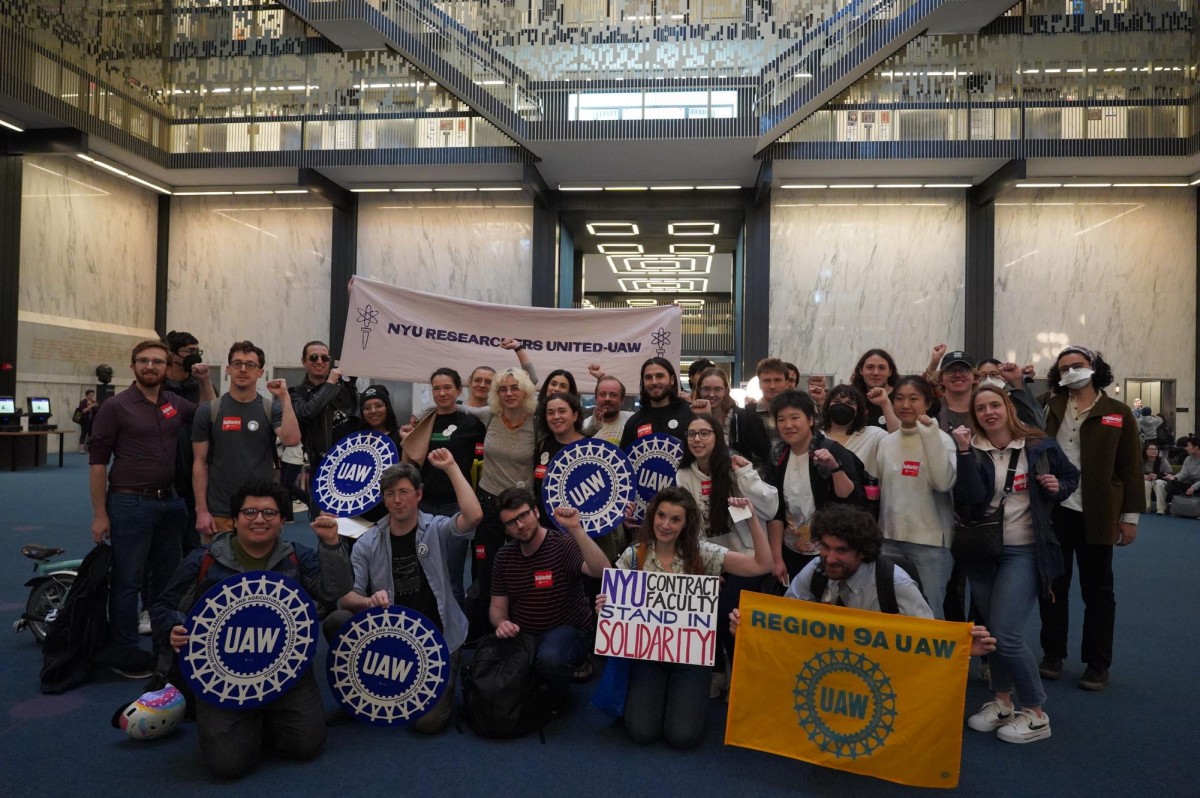 People are kneeling and standing on blue carpeting, a few holding blue and white signs that read ‘U.A.W.’ There is a white banner behind them that reads ‘N.Y.U. Researchers United-UAW,’ and a sign held by a woman kneeling in the front that reads, ‘N.Y.U. Contract Faculty Stand in Solidarity!’
