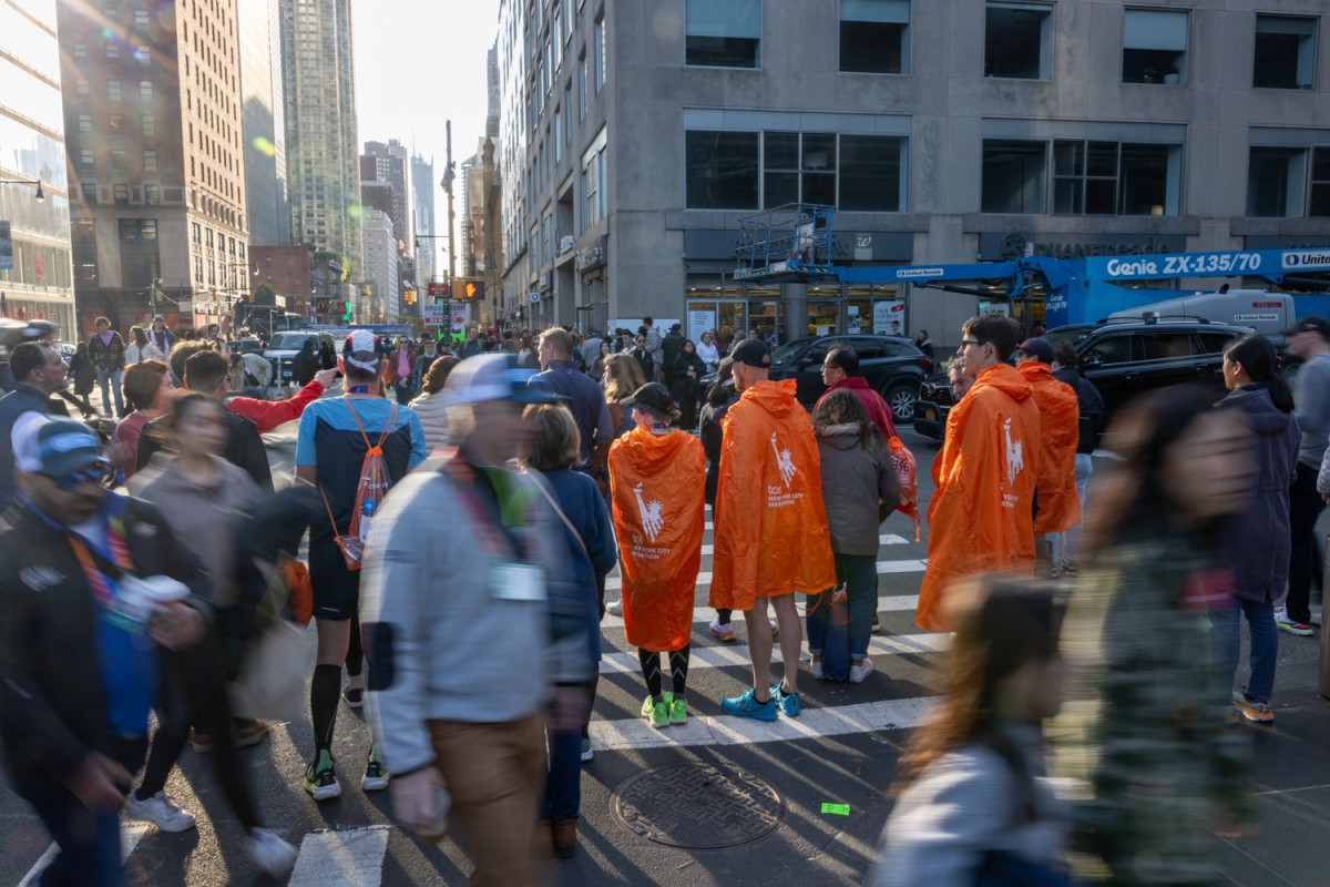 Four people in orange runners' ponchos stand at a crosswalk amidst a crowd of people.