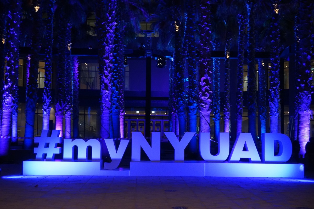 A sign which reads #myNYUAD lit by purple light sitting in front of a bunch of trees.