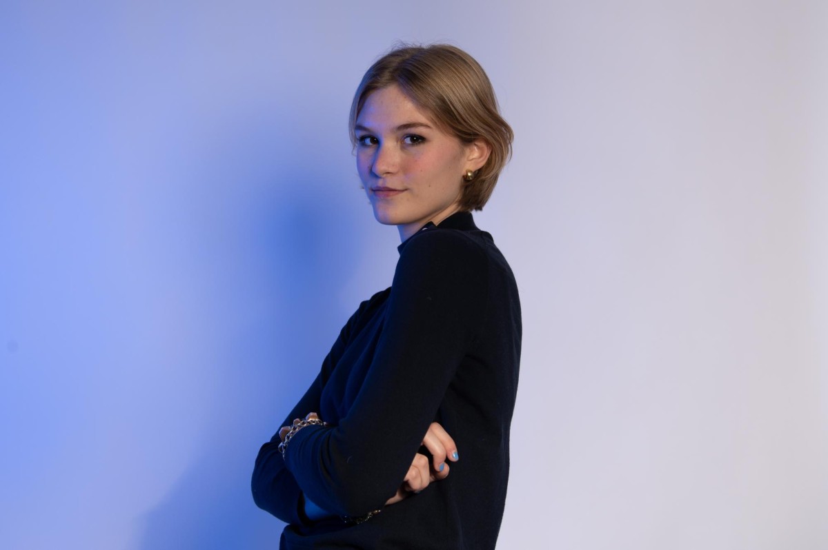 A woman with blond hair wearing a black shirt with her arms crossed across her chest. She has silver rings on her hands and is standing in front of a wall lit with blue lighting.