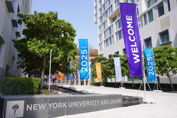 A stone sign which reads "NEW YORK UNIVERSITY ABU DHABI" stands in front of a long walkway with trees on both sides of it lined with banners reading "WELCOME" and "CLASS OF 2025" and "CLASS OF 2024" and "CLASS OF 2023"