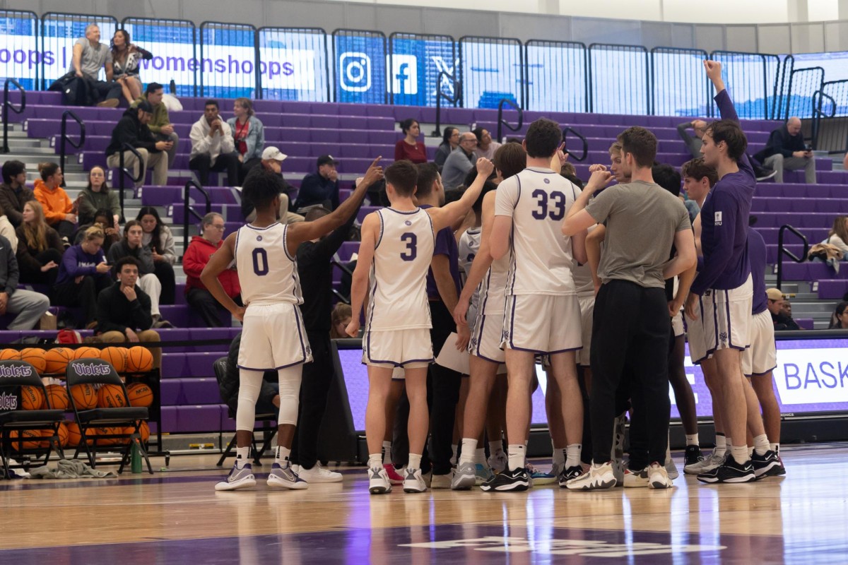 Men wearing white jerseys with purple numbers on them stand in a huddle with their arms raised in the center. Behind them are purple bleachers with people sitting on them.