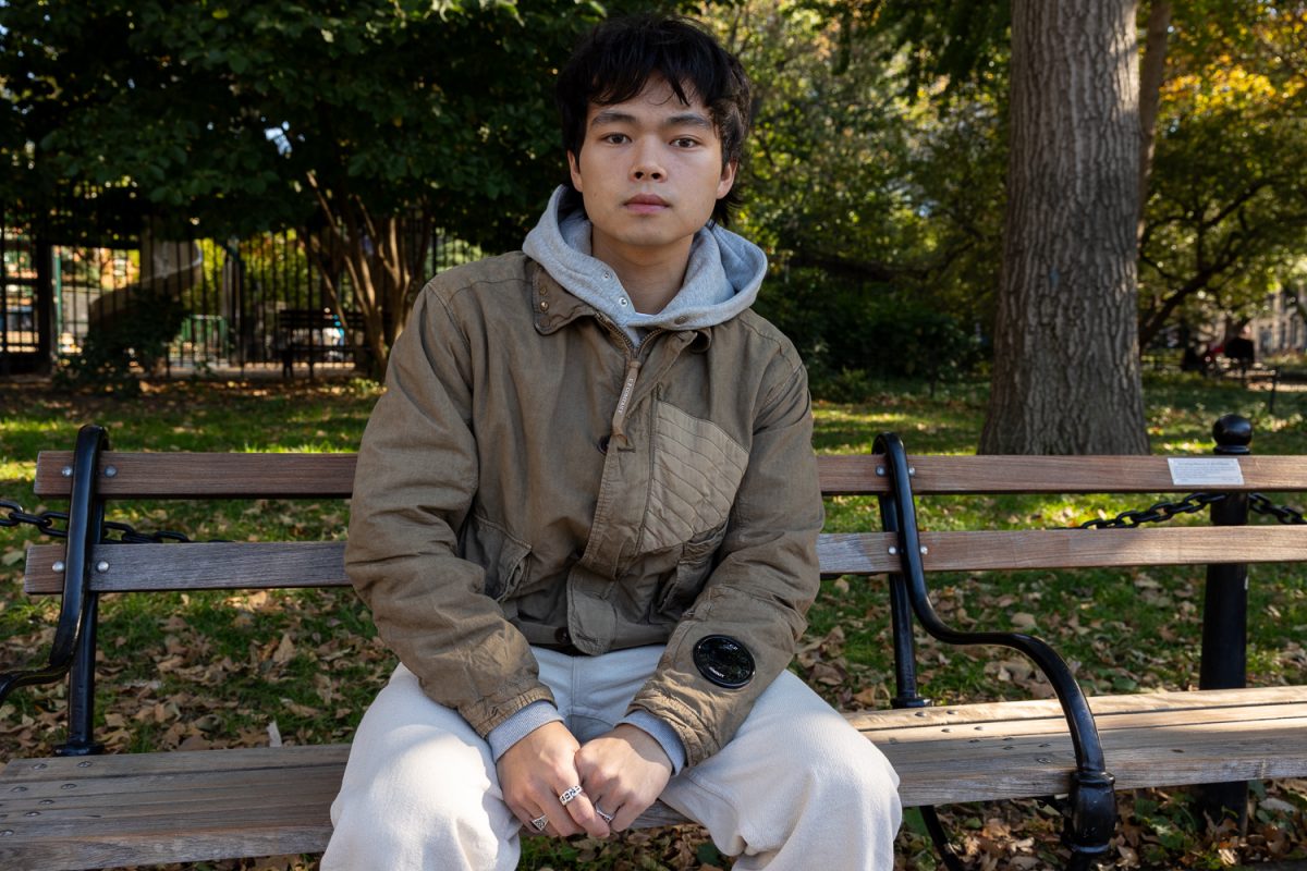 There is a man sitting on a bench in a park. He is wearing a brown jacket and faded white pants.