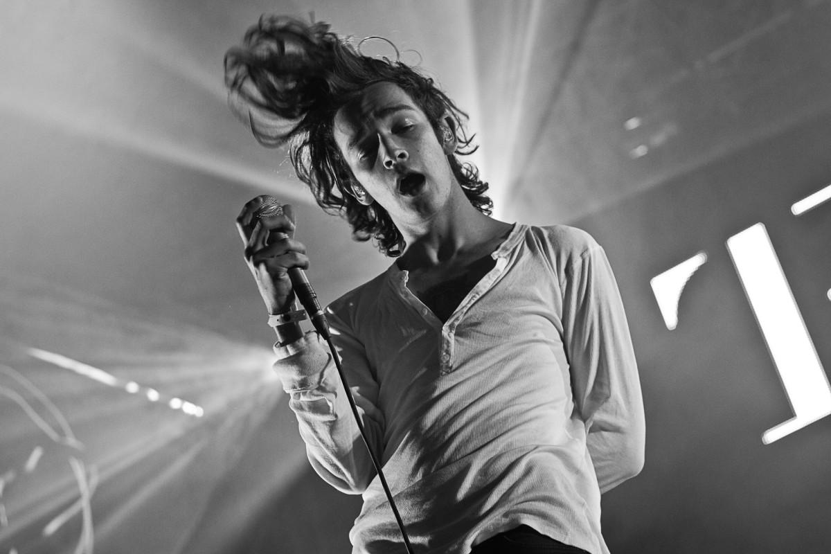 Black-and-white+close-up+photo+of+a+Matty+Healy+holding+a+microphone+in+one+hand+with+the+other+hand+behind+his+back+on+stage.