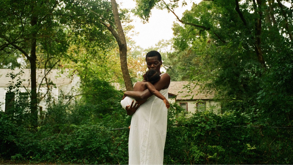 A person in a white dress stands holding a baby in a diaper. They are standing in the middle of green trees and foliage with a few houses in the background.