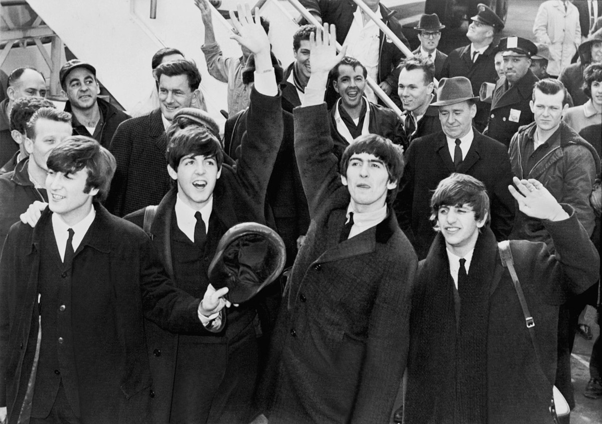 A black-and-white photo of the four members of The Beatles. They are all wearing suits. They appear to be raising their arms and waving. There are people behind them.