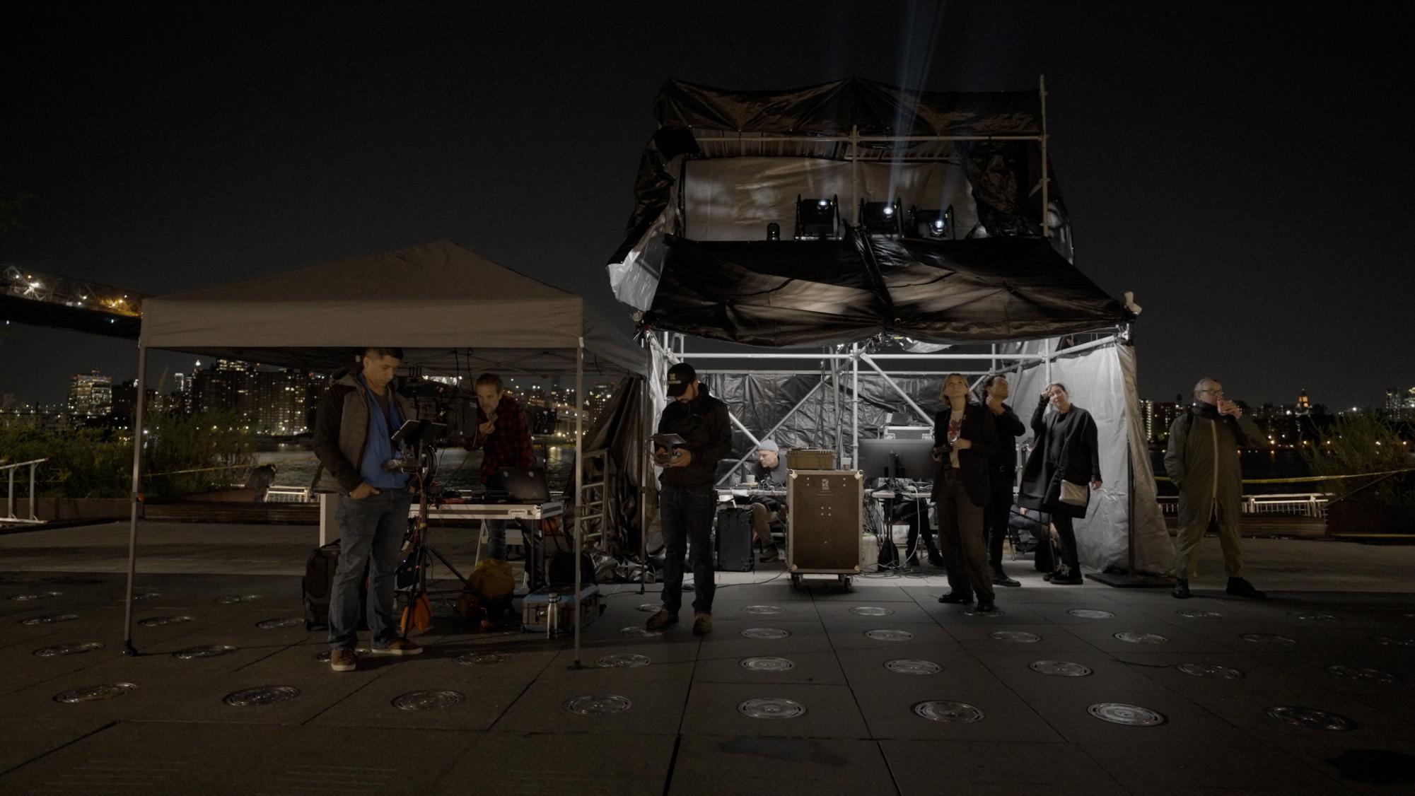 People stand outside at night in front of a set, with multiple tents and other equipment behind them.
