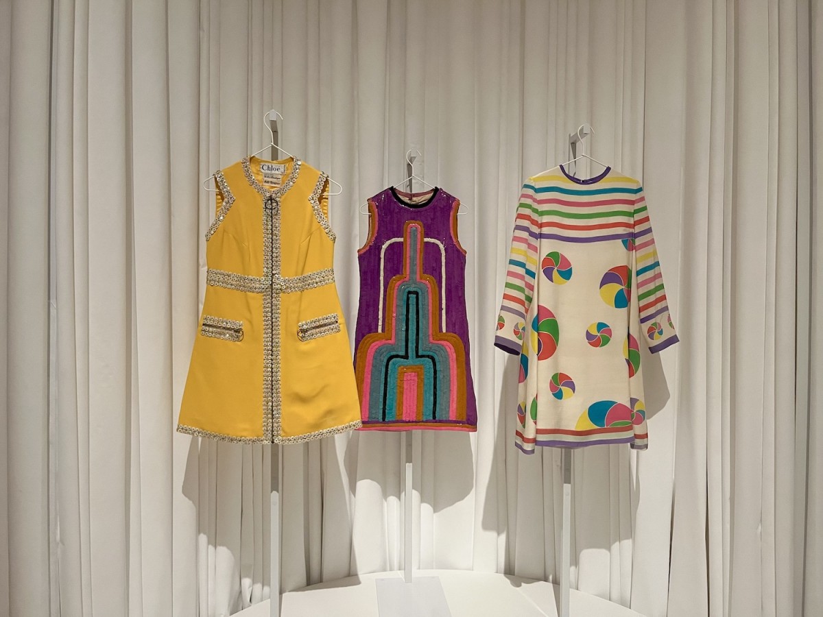 Three colorful dresses are hanging on white hangers. The dress on the left is yellow with silver beads, the one in the middle is purple, pink, orange, and blue, and the one on the right is cream-colored with colorful stripes and circles.