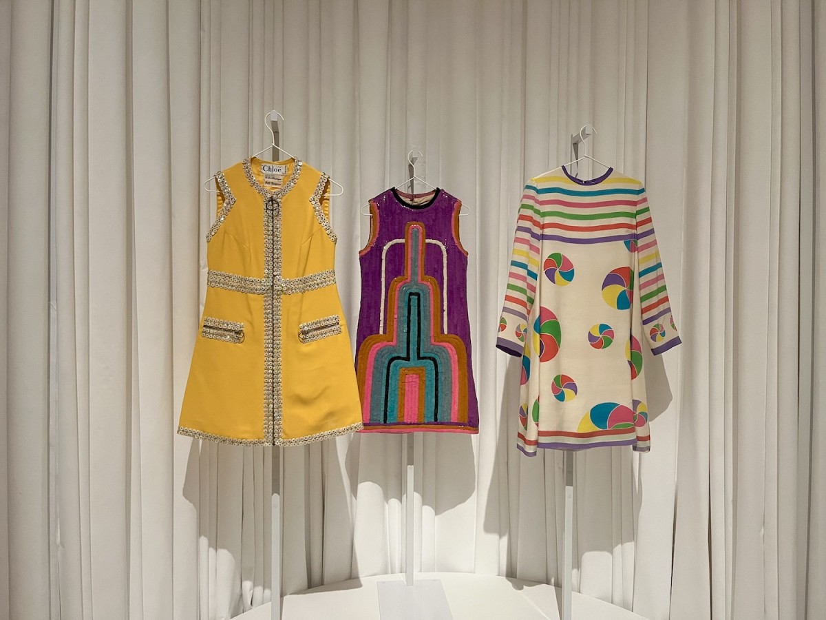 Three+colorful+dresses+are+hanging+on+white+hangers.+The+dress+on+the+left+is+yellow+with+silver+beads%2C+the+one+in+the+middle+is+purple%2C+pink%2C+orange%2C+and+blue%2C+and+the+one+on+the+right+is+cream-colored+with+colorful+stripes+and+circles.