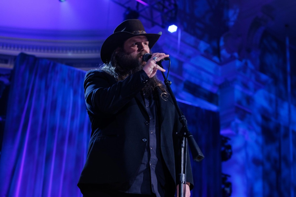 Chris Stapleton wearing a cowboy hat and a black coat while holding a microphone on a stand to his mouth. The stage behind him is bathed in blue light.