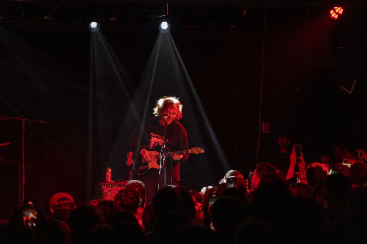 Two beams of light from a spotlight around a man covered in red light standing on a stage with a guitar. In front of him stands a crowd of people.
