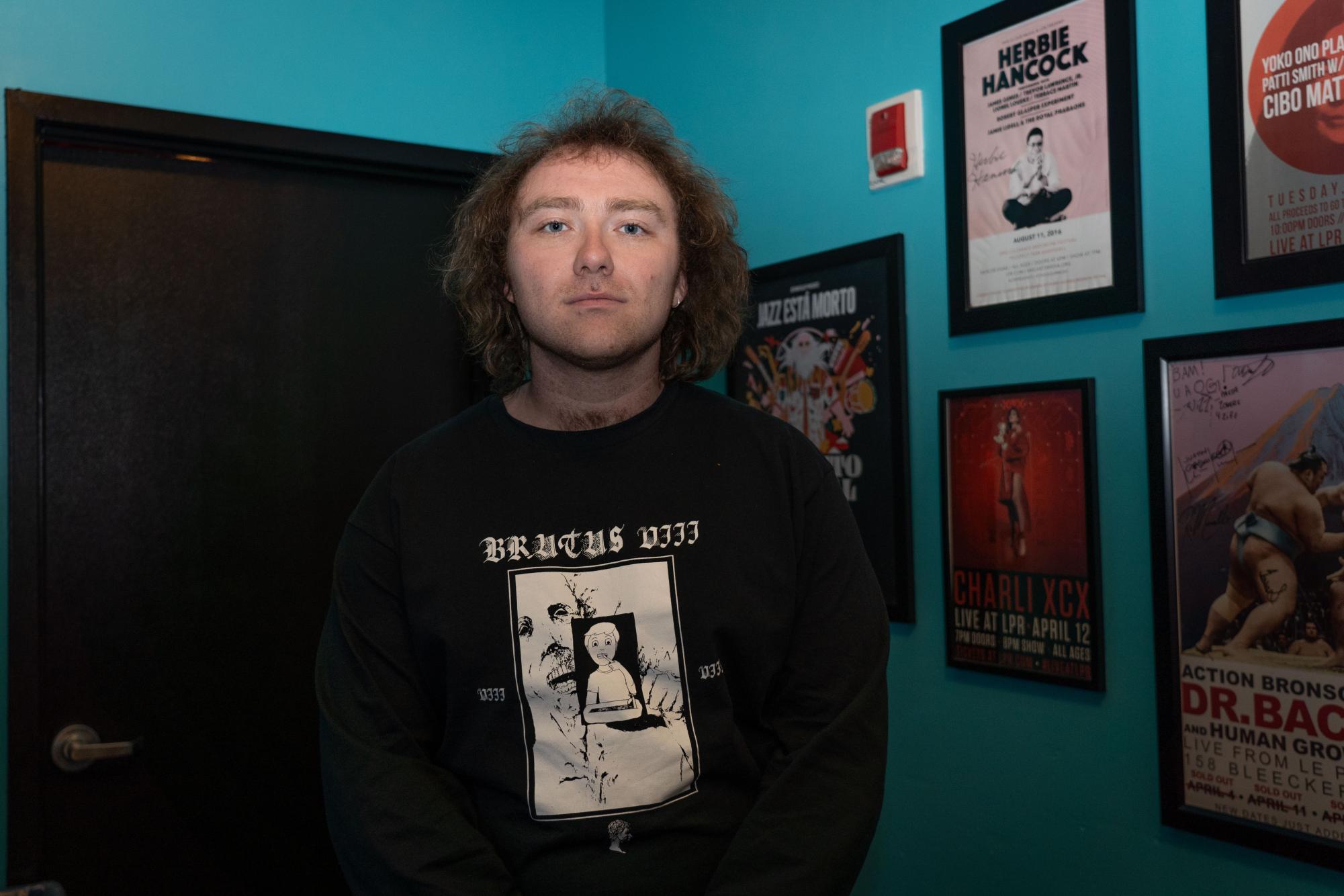 A man in a black t-shirt with the words “BRUTUS” on it stands in a turquoise room with