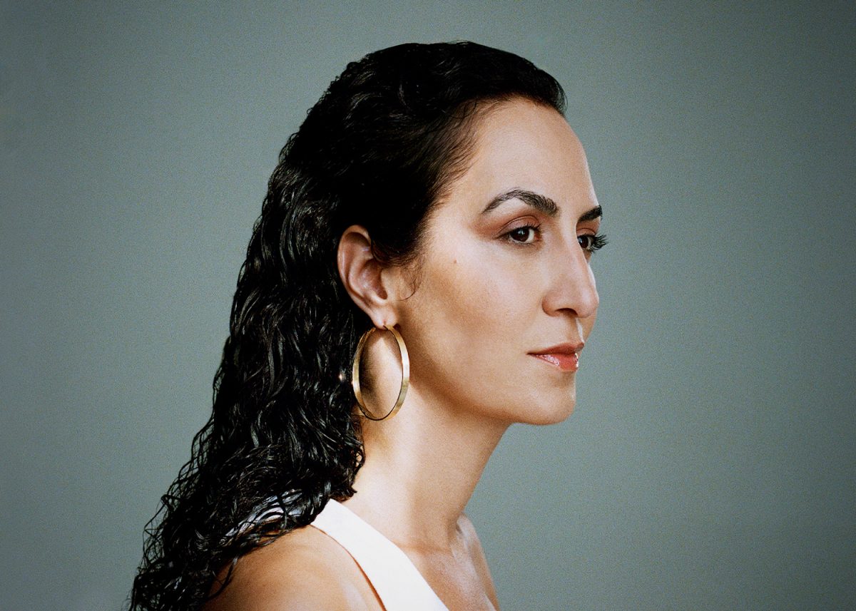 The side profile of a woman with long, dark brown hair. She is wearing a white top and golden hoop earrings.