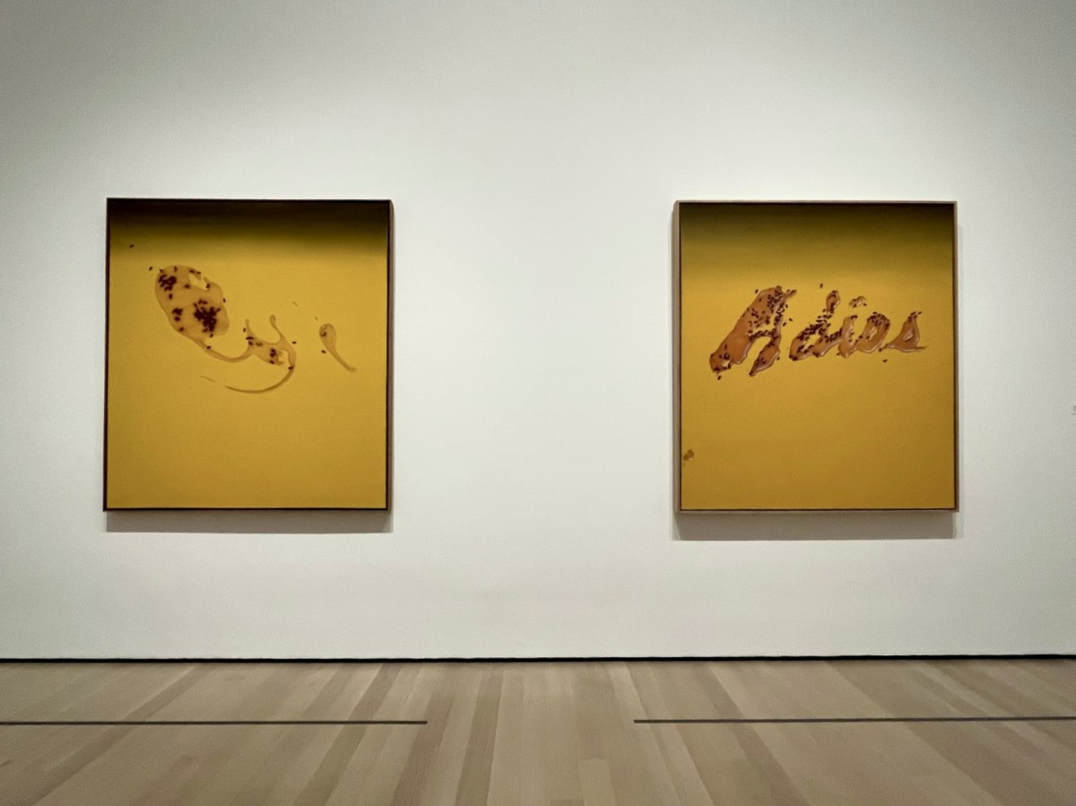 Two art prints hang on a white wall. They are both mustard yellow with the print on the right having the word “Adios”