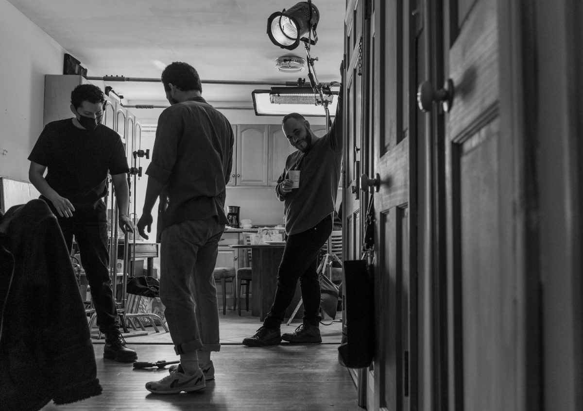 A black-and-white photo of a man holding a cup and resting his arm on a set of doors faces the camera. Behind him is a kitchen set and to his left are two people working with film equipment.