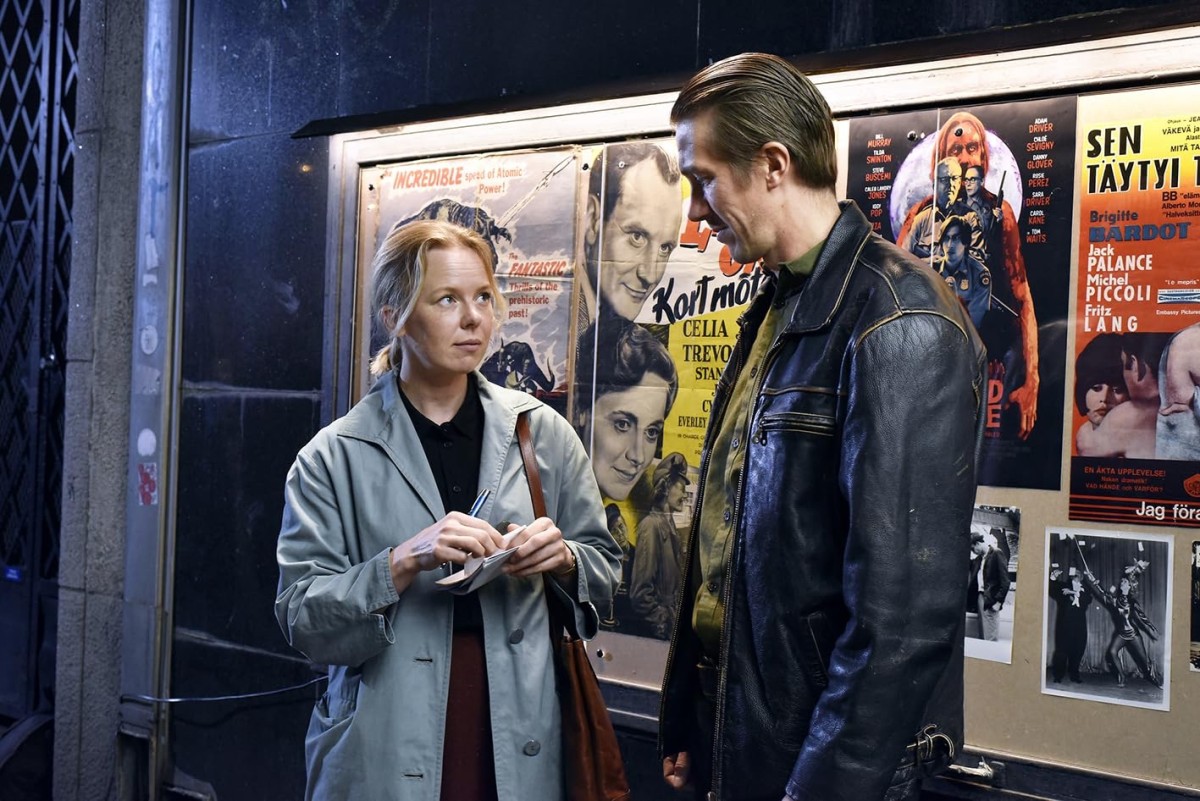 Two people are standing in front of a bulletin board with movie posters. They are looking at each other. One of them is wearing a gray jacket and the other one is wearing a black leather jacket.