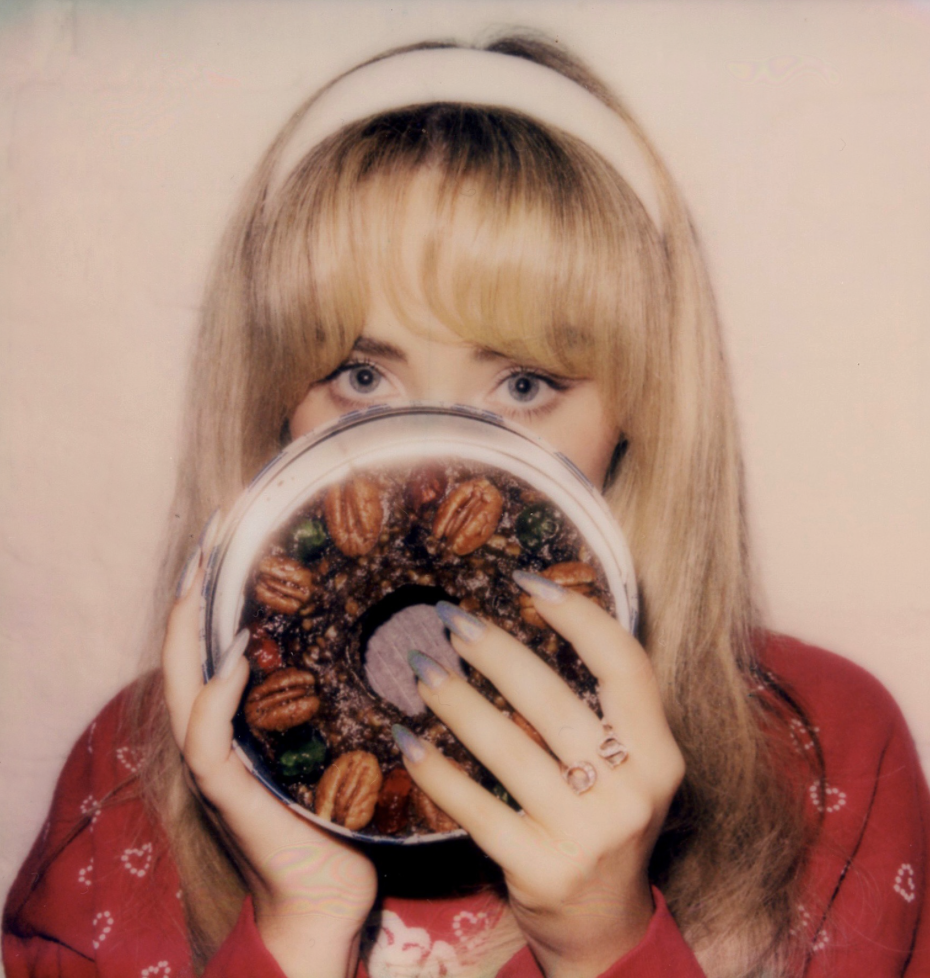 A girl with blond hair is holding a fruitcake in front of her face. She has a red top and white headband.