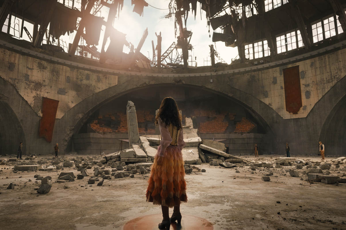 Rachel Zegler dressed as her character Lucy Gray Baird faces an area with rubble and a rooftop with a gaping hole in the middle.