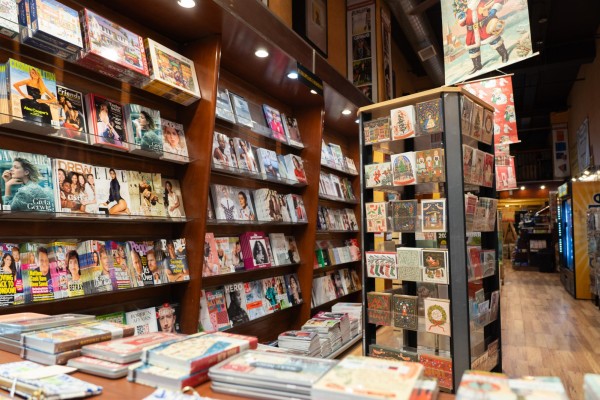 The interior of a magazine shop, where magazines are placed on a bookshelf by the wall and on a shorter shelf near the entrance. A rotating shelf with postcards on it is placed in the middle of the space.