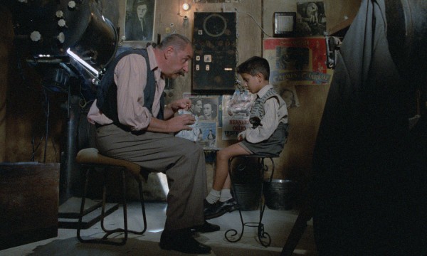 A still from the movie “Cinema Paradiso.” A man and a young boy sit across from each other in a dimly lit room.