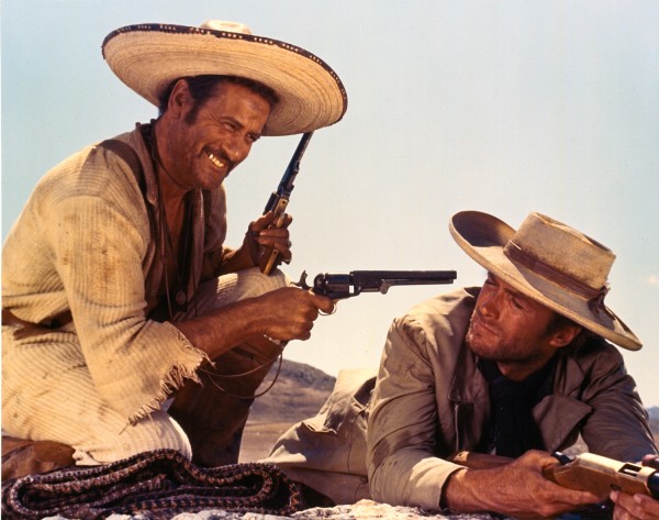 A still from the movie “he Good, The Bad and The Ugly.” There are two men — the man on the right lays on the ground propped up on his elbows and wears a hat. The man on the left kneels and playfully points a gun at the other man.