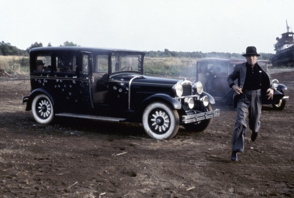 A still from the movie “Once Upon a Time in America.” A man dressed in a suit and top hat runs in front of a black car on a dirt path by a grass field.