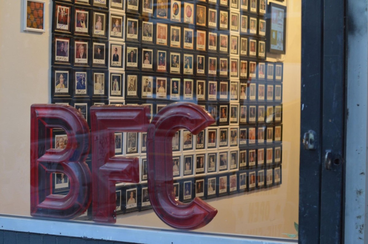 A storefront features a large red sign on the window that reads “B.F.C” Behind this there is a grid of autographed Polaroid photos on a wall.