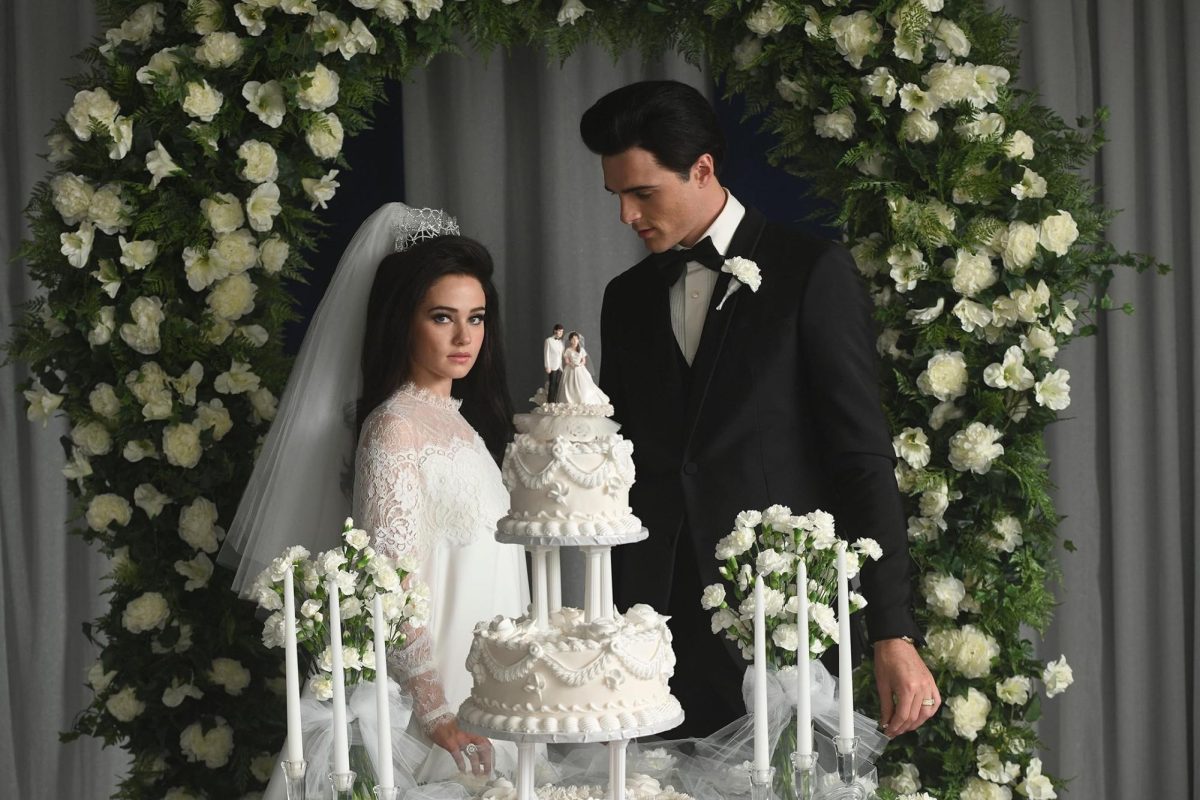 A bride and groom, both with dark brown hair, are standing behind a wedding cake, with bride and groom figures on top. There are white candles and white flowers on two sides of the cake, as well as a wreath with white flowers behind them.