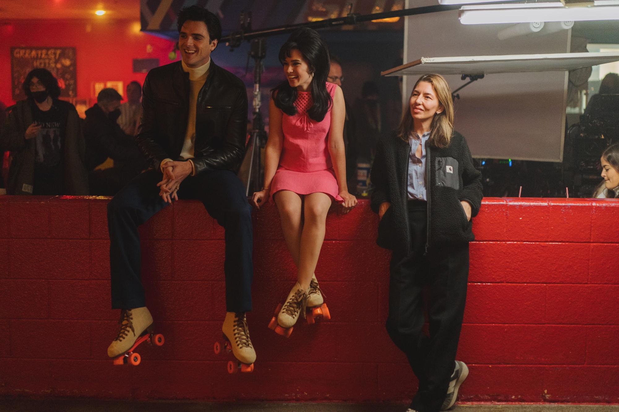 Jacob Elordi and Cailee Spaeny as their characters in “Priscilla” sit on a red wall wearing roller skates. Sofia Coppola stands next to them leaning against the wall.