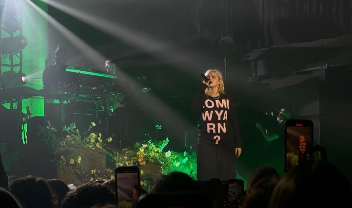 A woman with blond hair and a black shirt which reads “L.O.M.L W.Y.A. R.N. ?” stands on a stage with a microphone under green lights. In front of her below is a crowd of people holding up cell phones.