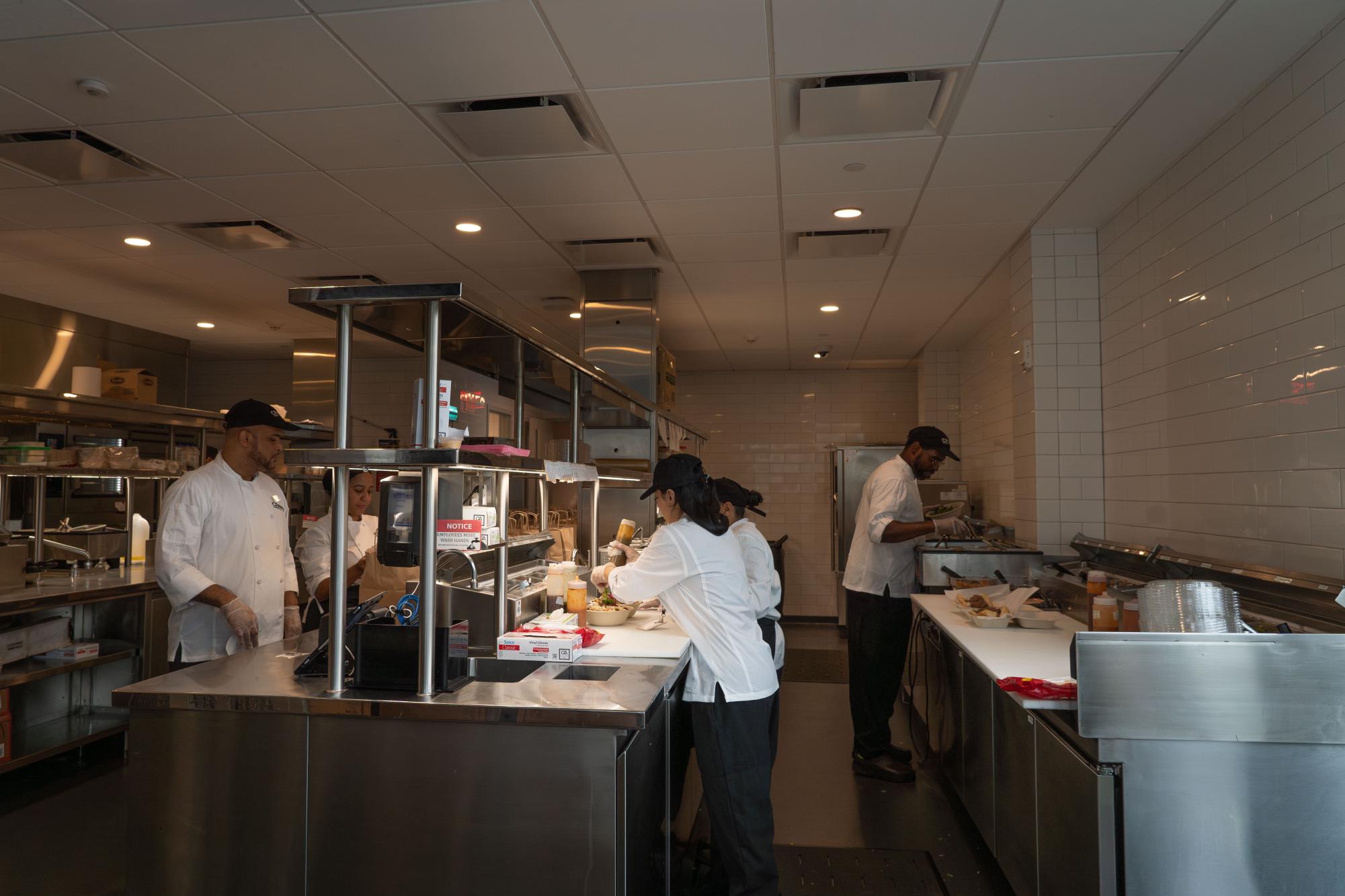 A large kitchen with various kitchen appliances. There are people wearing white coats, black pants, and black hats preparing food at various stations around the kitchen.