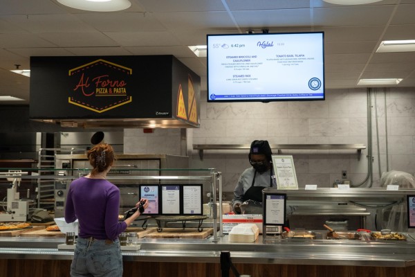 A woman wearing a purple shirt and light blue jeans stands in front of a food stall, while a staff from N.Y.U. Eats stands behind it. Above the stall is a television screen displaying a menu of Halal food offered at Kimmel Marketplace.