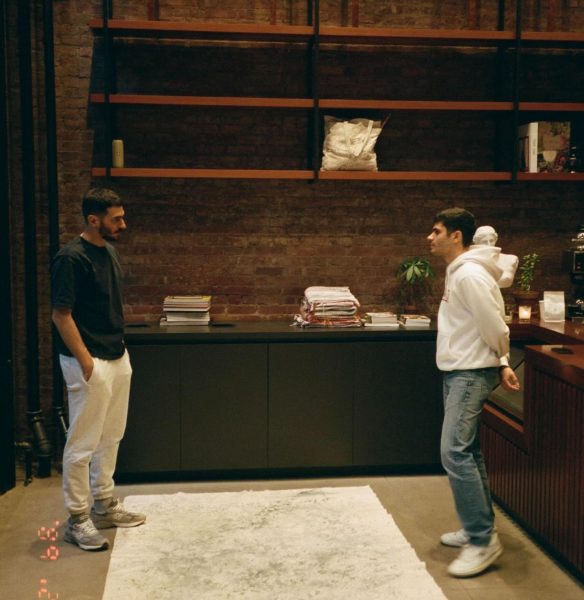 Two people are standing and looking at each other. One of them is wearing a black t-shirt and gray pants, and the other is wearing a white hoodie and jeans. There is a brick wall with brown shelves and black cupboards behind them.
