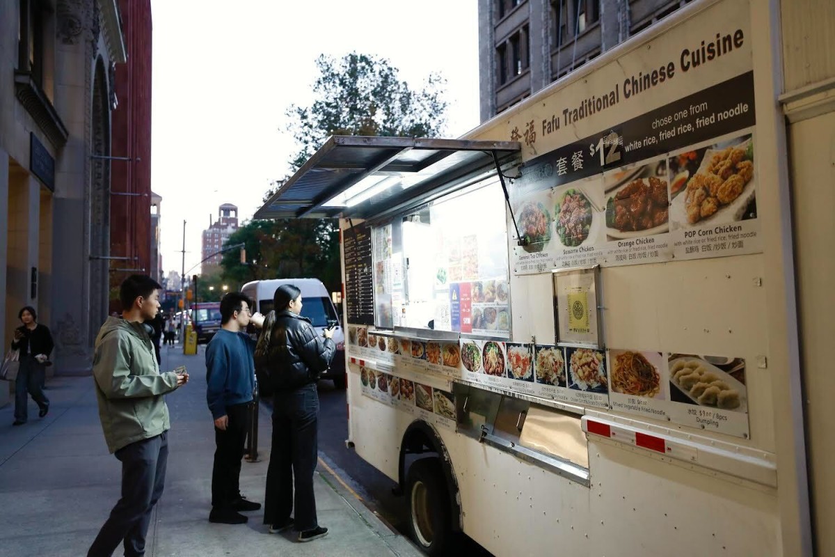 A white food truck that reads on the side “Fafu Traditional Chinese Cuisine” and photos of different menu items posted on its exterior parks outside N.Y.U.’s Stern School of Business. Three people are standing outside the food truck waiting for their food.
