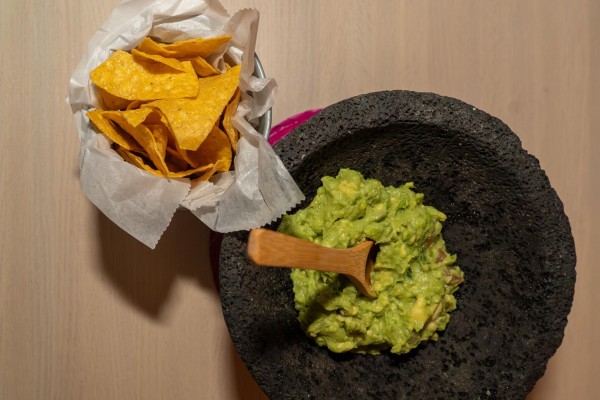 A scoop of guacamole is pictured inside of a molcajete with a bowl of tortilla chips to its upper left.