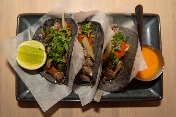 Three beef tacos placed side by side on a black, rectangular plate. A slice of lime and a side of an orange sauce were placed on the plate.
