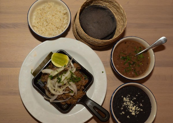A white plate with a black iron pan placed on it. On the iron pan, a slice of lime and pieces of white onions are placed on top of a serving of beef. Four bowls are placed around the plate, with white rice, black taco shells, beans and black sauce placed in each of them.
