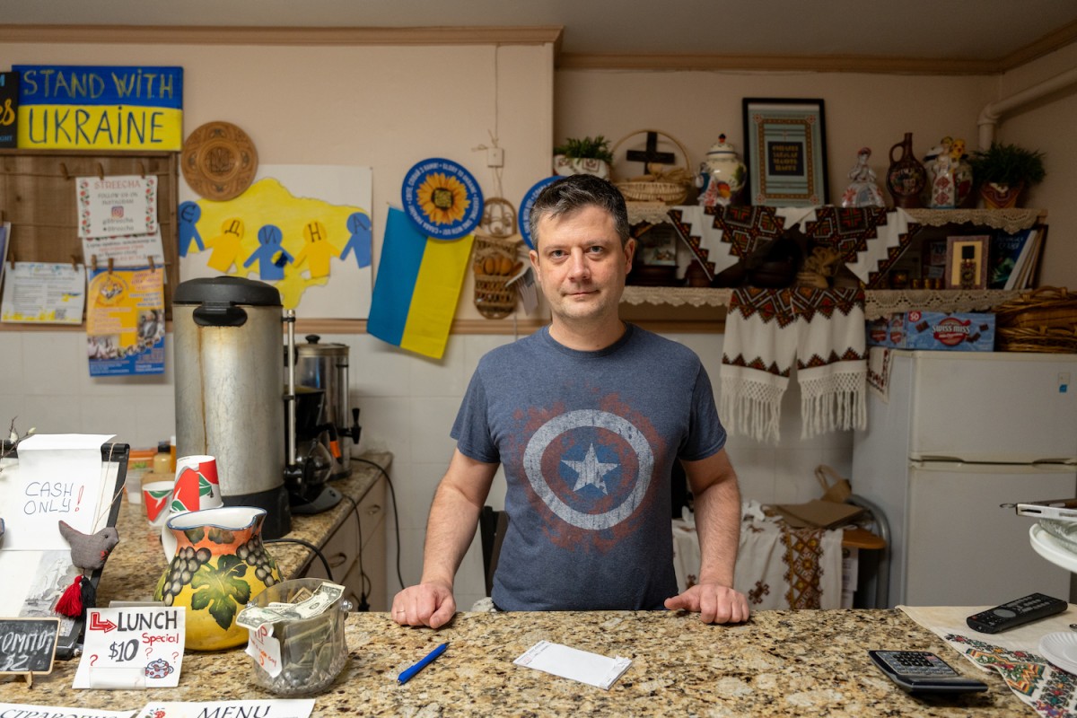 A+man+wearing+a+dark+blue+T-shirt+with+a+Captain+America+shield+image+on+it+stands+behind+a+restaurant+counter.+Behind+him+is+a+wall+decorated+by+Ukrainian+flags%2C+fabrics%2C+and+pro-Ukrainian+signs+and+flyers.