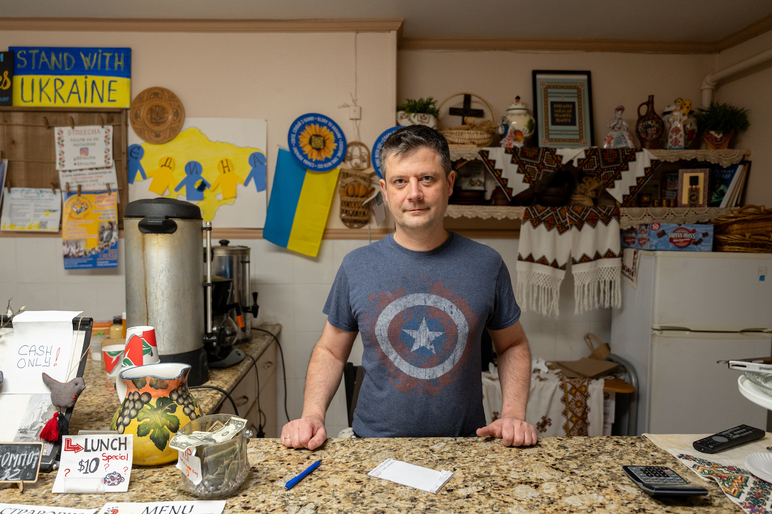 A man wearing a dark blue T-shirt with a Captain America shield image on it stands behind a restaurant counter. Behind him is a wall decorated by Ukrainian flags, fabrics, and pro-Ukrainian signs and flyers.