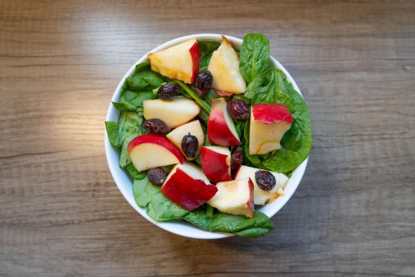 A white bowl with spinach, cranberries and apples placed on a table.